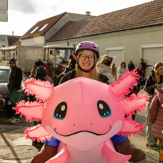 Eine Frau in einem pinken Drachen-Mascot-Anzug posiert für ein Foto auf der Straße. Hinter ihr gehen mehrere Leute, einige tragen Helme und Brillen. In der Nähe steht ein Auto und ein Gebäude.