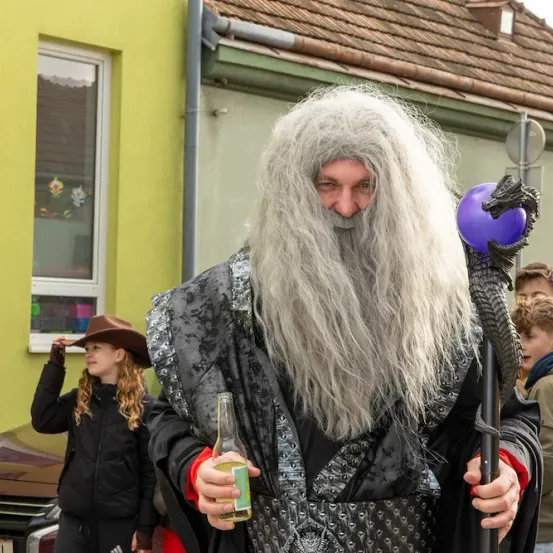 Ein Mann, der als Gandalf gekleidet ist, mit einem violetten Orb-Stab und einer Bierflasche, steht vor einem gelben Gebäude. Ein Mädchen und zwei Jungen beobachten ihn.