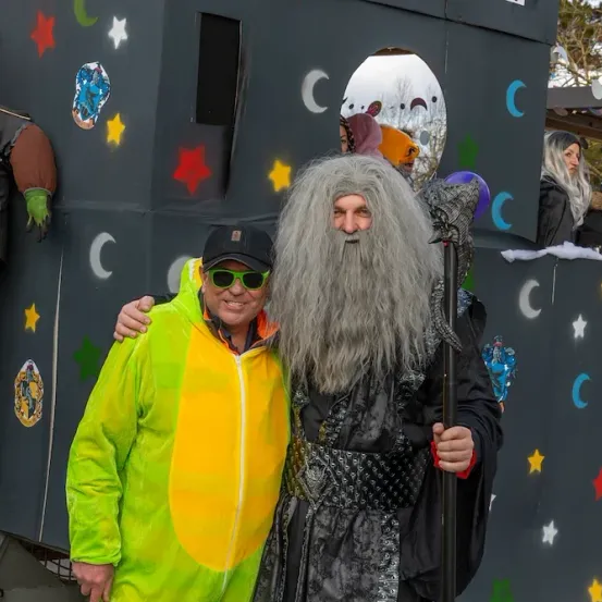 Zwei Männer in Kostümen, einer mit langem Bart und einem Stab, stehen vor einer dekorierten Wand.