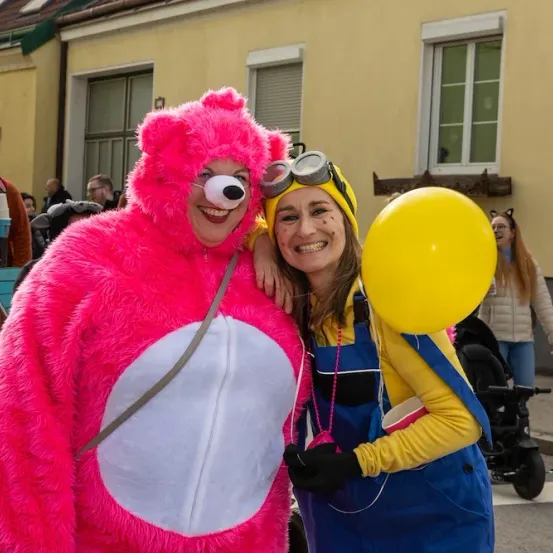 Ein rosafarbener Bären-Mascot steht neben einer lächelnden Frau in einem gelben Kostüm, beide halten einen gelben Ballon, mit einem Gebäude und einer Menge im Hintergrund.