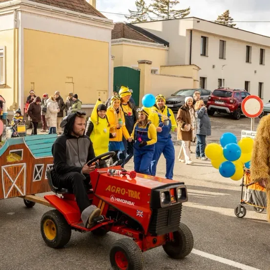 Eine Gruppe von Menschen in Kostümen fährt einen roten Traktor, während andere in Kostümen dahinter gehen. Sie halten Luftballons und stehen in einem Umzug vor Gebäuden.