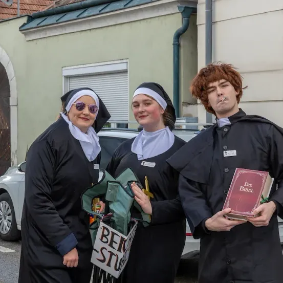 Drei Personen, verkleidet als Mönche und Nonnen, stehen vor einem Gebäude. Einer hält ein Buch, ein anderer ein Schild. Ein Auto ist dahinter geparkt.