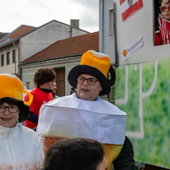 Zwei Menschen in Bierkrug-Kostümen stehen vor einem geschmückten Lastwagen. Dahinter sind weitere Menschen in verschiedenen Kostümen zu sehen.