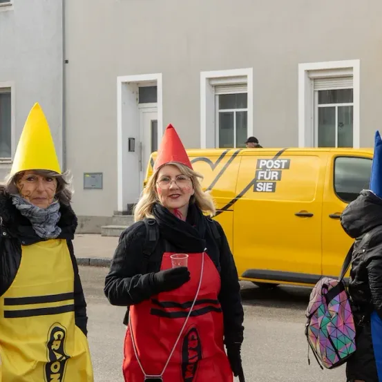 Drei Personen in Buntstift-Kostümen stehen vor einem gelben Van mit der Aufschrift 'Post für Sie'. Die Frau in der Mitte hält einen Becher. Ein Gebäude mit weißen Fensterrahmen ist im Hintergrund.