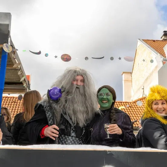 Drei Menschen in Kostümen stehen auf einem Festwagen. Einer hält einen lila Luftballon, ein anderer ein Weinglas und ein dritter hält einen blauen Stab.