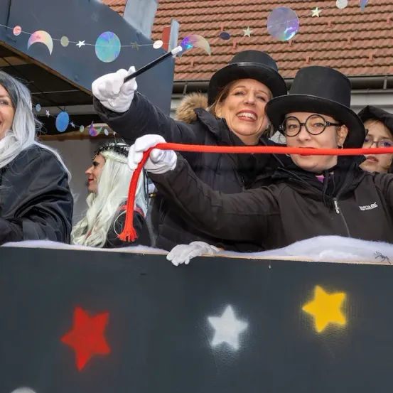 Vier Frauen in Kostümen, darunter Hexen und Magierinnen, stehen auf einem Wagen und halten ein rotes Band. Sie lächeln und scheinen ein Ereignis zu feiern.