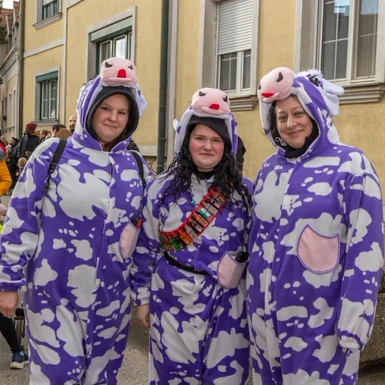 Drei Frauen in lila Kuhkostümen stehen vor einem Gebäude. Zwei von ihnen haben Kuhohren und eine trägt einen Gürtel mit mehreren Gegenständen. Sie lächeln für ein Foto.