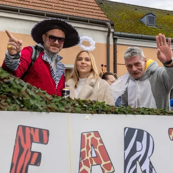 Drei Menschen in Festkleidung winken von einem Wagen mit einem Schild, das 'F A R M' sagt. Dahinter ein Gebäude mit moosbewachsenem Dach und Schornstein.