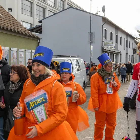 Eine Gruppe von Menschen in orangefarbenen Kostümen geht auf einer Straße. Eine Frau hält eine Flasche Aperol. Ein Mann trägt ein rotes und weißes Outfit. Sie stehen vor einem Gebäude.