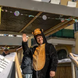 Ein Mann in einem Bärenkostüm winkt vor der Kamera, während er unter einem Baldachin mit dekorativen Lichtern steht. Dahinter hält eine Person ein Schild. Gebäude und ein Balkon sind sichtbar.