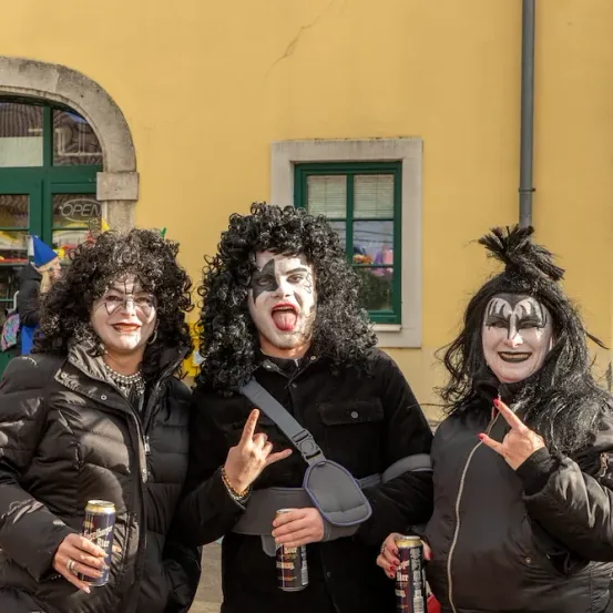 Drei Personen in KISS-Bandkostümen posieren mit Bierdosen vor einem gelben Gebäude.