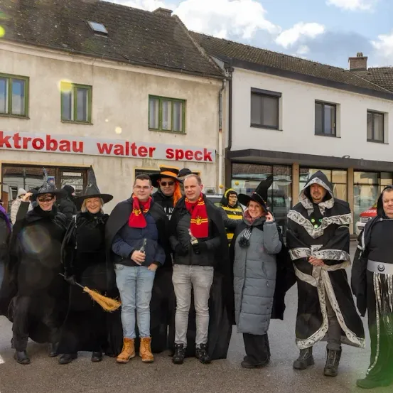 Eine Gruppe von Menschen in Kostümen steht vor einem Gebäude mit der Aufschrift Elektrobau Walter Speck. Sie lächeln für ein Foto an einem bewölkten Tag.