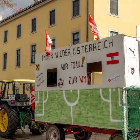 Ein Anhänger mit einem Fußballfeld-Design, mit dem Slogan 'Immer wieder Österreich' und Fahnen, ist vor einem Gebäude geparkt.