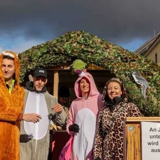 Vier Personen in Tierkostümen stehen vor einer getarnten Struktur und lächeln. Ein Schild warnt, dass Kinder unter 16 Jahren nicht zugelassen sind.