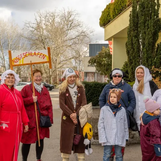 Eine Gruppe von Erwachsenen und Kindern in Kostümen, eine mit einem Schild 'AUSGESCHLAFEN', stehen vor einem Gebäude mit Bäumen im Hintergrund.