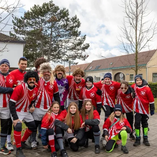 Eine Gruppe von Menschen in roten und weißen Uniformen posiert für ein Foto, einige mit Gesichtsbemalung, vor einem Gebäude mit einem großen Baum.
