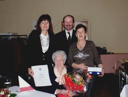 Eine Gruppe von vier Personen steht hinter einem Tisch mit einem Blumenstrauß, einem Zertifikat und einem Geschenk. Sie scheinen ein Ereignis zu feiern.