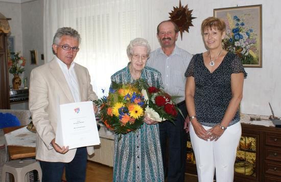 Eine ältere Frau mit Blumen steht zwischen zwei Männern, einer hält ein Zertifikat. Sie sind in einem Raum mit weißen Wänden und einem Blumenbild.