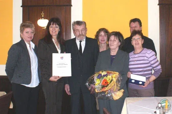 Eine Gruppe von Menschen steht in einem Raum und hält Urkunden und Blumen, möglicherweise für eine Preisverleihung.