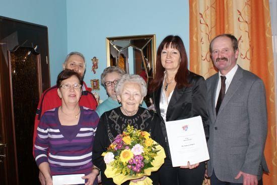 Eine Gruppe von Menschen steht in einem Raum, hält Urkunden und Blumen. Die Frau in der Mitte hält einen großen Blumenstrauß.