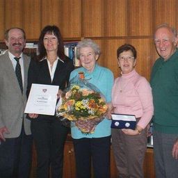 Eine Gruppe von Menschen steht zusammen, zwei halten Blumen und Zertifikate. Sie befinden sich in einem Raum mit Holzschränken und Büchern.