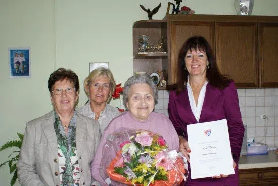 Eine Frau mit einem Zertifikat steht mit drei anderen, eine hält einen Blumenstrauß, in einer Küche. Dahinter steht ein Holzregal mit verschiedenen Gegenständen.
