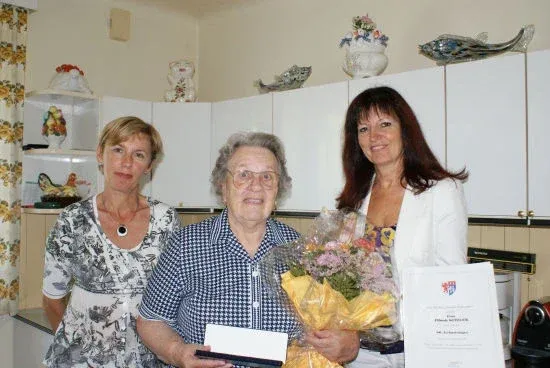 Drei Frauen stehen in einer Küche, wobei die mittlere Frau einen Blumenstrauß und ein Zertifikat hält, während die anderen Geschenke halten. Hinter ihnen befinden sich weiße Schränke und dekorative Gegenstände.
