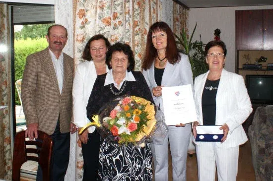 Fünf Personen posieren für ein Foto im Innenraum. Eine Frau hält Blumen und ein Zertifikat. Eine andere Frau hält eine Schachtel.