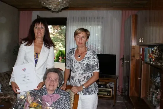 Eine Frau sitzt in einem Stuhl, hält einen Blumenstrauß und ein Zertifikat in der Hand, mit zwei Frauen, die neben ihr stehen, eine hält ein Zertifikat. Dahinter steht ein Sofa, ein Fernseher und ein Fenster mit weißen Vorhängen.