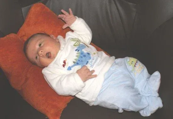Ein Baby liegt auf einer Couch mit einem orangefarbenen Kissen und trägt ein weißes Langarmshirt mit Dinosaurier-Aufdruck.