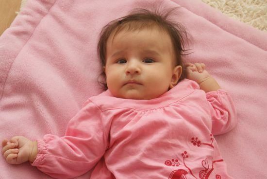 Ein Babymädchen mit braunen Haaren liegt auf einer rosa Decke. Sie trägt ein rosa Langarmshirt. Ihre Hände sind erhoben, und ihre Augen sind weit geöffnet.
