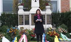 Eine Frau in einem schwarzen Mantel steht vor einem Denkmal, hält ein Papier in der Hand, mit Blumen und Bändern drumherum.
