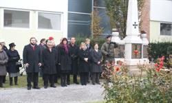 Eine Gruppe von Menschen in Mänteln steht vor einem Denkmal mit Blumen und einem Kreuz. Dahinter befindet sich ein Gebäude mit Glasfenstern.