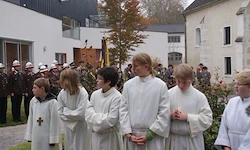 Eine Gruppe von Kindern in weißen Gewändern steht zusammen vor einem Gebäude, hinter dem mehrere Menschen in Uniform stehen.