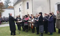 Ein Chor steht vor einer Kirche und singt. Der Dirigent in einem schwarzen Mantel hält ein Notenpult. Die Chormitglieder sind in Winterkleidung gekleidet.
