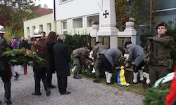 Eine Gruppe von Menschen, darunter einige in Militäruniformen, stehen um ein Denkmal herum. Sie legen Kränze und Blumen darauf, einige tragen Mützen und Stiefel.