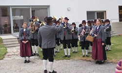 Eine Blaskapelle in traditionellen Uniformen spielt auf einem Kiesweg vor einem Gebäude.