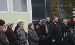 Eine Gruppe von Menschen, die warm gekleidet sind, steht vor einem Gebäude, manche mit gefalteten Händen. Ein Mann mit Bart und roter Krawatte steht vorne.