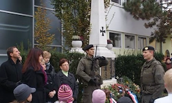 Eine Gruppe von Menschen in Winterkleidung steht vor einem Denkmal, mit zwei Soldaten in Uniform in der Nähe und einem Gebäude mit einem Kreuz im Hintergrund.