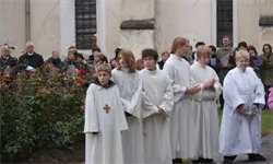 Eine Gruppe von Menschen in weißen Gewändern steht vor einer Kirche, mit Blumen und einer Menschenmenge im Hintergrund.
