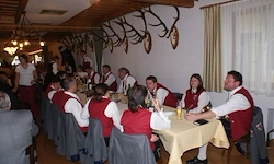 Eine Gruppe von Menschen in roten Westen sitzt an einem Tisch in einem Raum mit Holzbalken, Hirschgeweihen und einer Laterne. Sie essen eine Mahlzeit.