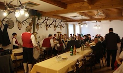 Ein Speisesaal mit Holzbalken und einer Kronleuchter. Eine Band spielt, während eine Menge isst. Ein Mann mit einer Trommel steht vor einem Tisch mit Flaschen und Tellern.