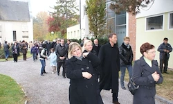 Eine Gruppe von Menschen in Wintermänteln geht auf einem Kiesweg vor einem Gebäude entlang, einige schauen in die Kamera.