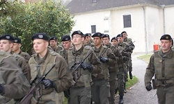 Eine Gruppe von Militärangehörigen in Tarnuniformen marschiert in Formation einen Pfad hinunter, angeführt von einem Kommandanten. Bäume und ein Gebäude sind im Hintergrund zu sehen.