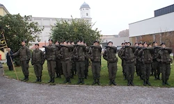 Eine Gruppe von Soldaten in Uniform steht in Formation auf dem Rasen vor einem Gebäude. Sie halten Gewehre und scheinen zu salutieren.