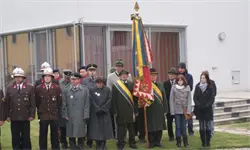 Eine Gruppe von Menschen in Uniformen und Mänteln steht vor einem Gebäude und hält eine Flagge und ein Banner. Einige schauen in verschiedene Richtungen.