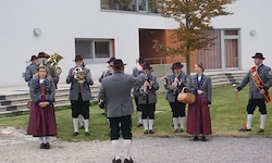 Eine Gruppe von Musikern in traditioneller Kleidung steht auf Gras vor einem Gebäude. Sie halten verschiedene Instrumente, darunter Blech- und Holzblasinstrumente. Die Szene spielt im Freien, mit Stufen und Fenstern im Hintergrund.