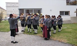 Eine Gruppe von Musikern in traditioneller Kleidung, einschließlich Hüten und Lederschuhen, spielen Blechblasinstrumente auf dem Rasen vor einem weißen Gebäude.