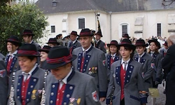 Eine Gruppe von Menschen in Uniform, mit Medaillen und Hüten, geht in einer Reihe vor einem Gebäude. Einige tragen Fahnen.