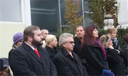 Eine Gruppe formell gekleideter Personen steht vor einem Gebäude, einige tragen Mützen und Schals. Die Szene ist im Freien mit Bäumen und einem Topfpflanzen.
